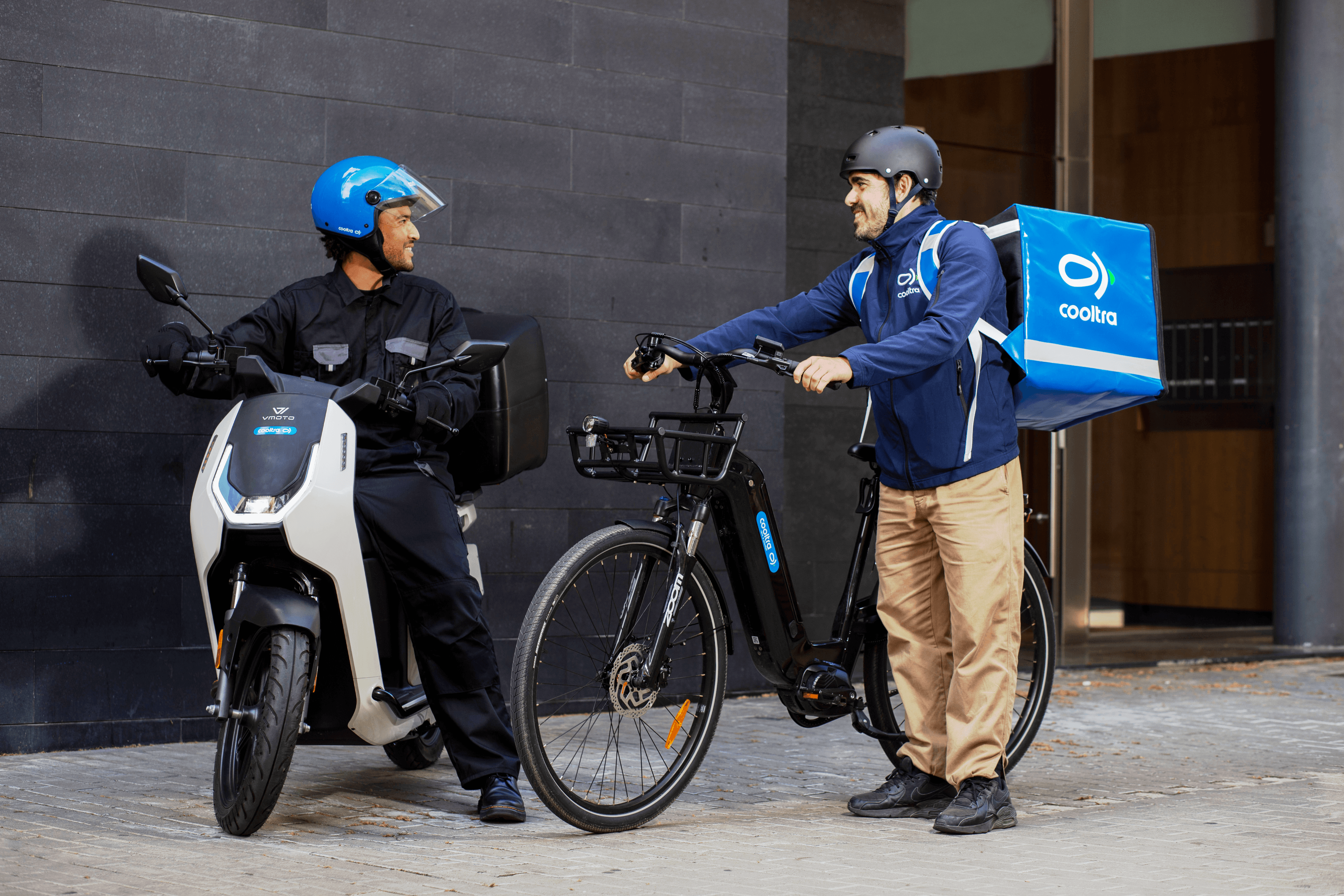 Mujer con casco sonriendo junto a su bicicleta de reparto bajo un pasillo con arcos.